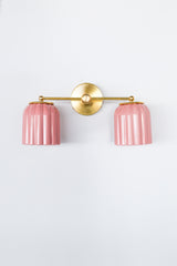 Front view of the Double Marcella Sconce featuring two glossy ribbed ceramic shades in soft blush pink on a brass fixture, a colorful ceramic wall sconce for bathrooms, traditional homes, and playful vintage inspired spaces, offering a dreamy pastel pop for color lovers and kids bathrooms