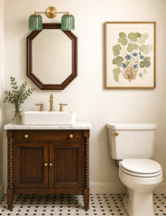 Neutral bathroom vanity with a double Marcella wall sconce in glossy jade green ceramic ribbed shades and warm brass hardware, mounted above an octagon wood mirror. Classic white vessel sink on a dark stained wood vanity, brass faucet and knobs, and black-and-white penny tile floor create a vintage inspired, grandmillennial feel. Botanical wall art and a simple glass vase add an organic touch. Elegant double bathroom light fixture for traditional, transitional, and cottage style interiors.