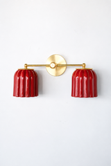 Handmade Double Marcella Sconce in jasper red glazed ceramic — two fluted, scalloped shades on a slim brass arm and round backplate. The glossy oxblood glaze pools richer and darker in the flutes and catches light on the raised ribs, so the color reads as a deep jewel-red that shifts from bright cherry to smoky garnet as it curves. Deco-inspired ribbing keeps the silhouette feminine and architectural, while brass warms the whole piece. This small-batch ceramic sconce makes a confident statement flanking a m