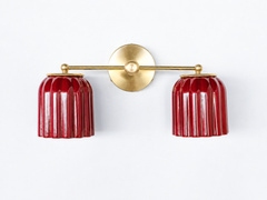 Handmade Double Marcella Sconce in jasper red glazed ceramic — two fluted, scalloped shades on a slim brass arm and round backplate. The glossy oxblood glaze pools richer and darker in the flutes and catches light on the raised ribs, so the color reads as a deep jewel-red that shifts from bright cherry to smoky garnet as it curves. Deco-inspired ribbing keeps the silhouette feminine and architectural, while brass warms the whole piece. This small-batch ceramic sconce makes a confident statement flanking a m