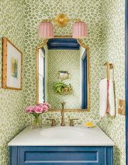 Vibrant powder room featuring the Double Marcella Sconce with ribbed pastel pink ceramic shades and a warm brass backplate, mounted above a marble vanity with navy cabinetry and brass faucet. The bathroom is styled with bold green spotted wallpaper, a gold-trimmed mirror, and fresh pink flowers, showcasing how handmade ceramic wall lighting by Sazerac Stitches adds playful Art Deco-inspired elegance and a pop of color to eclectic, modern interiors.