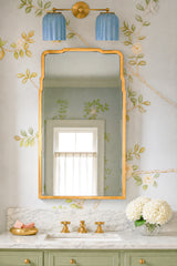 Grandmillennial bathroom vanity styled with a sky blue double Marcella wall sconce featuring glossy ribbed ceramic shades and warm brass hardware. Fixture is mounted above a curved-corner brass mirror on a marble countertop with brass widespread faucet, set against soft botanical floral wallpaper. Sage green vanity cabinetry and classic white sink create an airy, vintage-inspired look. Elegant double bathroom light for traditional, cottage, and transitional interiors, perfect for powder rooms and primary va