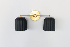 Handmade Double Marcella Sconce in unglazed matte black ceramic — two fluted, scalloped shades on a slim brass arm and round backplate. The bare, unglazed clay gives each shade a soft, stony matte surface with subtle handmade variation, while the Deco-inspired ribbing keeps the silhouette feminine and architectural at once. Black grounds the brass beautifully, so this small-batch ceramic sconce works as a graphic statement or a quiet anchor — flanking a mirror, framing a bed, or lining a hallway. The downwa