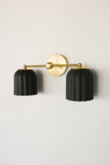 Handmade Double Marcella Sconce in unglazed matte black ceramic — two fluted, scalloped shades on a slim brass arm and round backplate. The bare, unglazed clay gives each shade a soft, stony matte surface with subtle handmade variation, while the Deco-inspired ribbing keeps the silhouette feminine and architectural at once. Black grounds the brass beautifully, so this small-batch ceramic sconce works as a graphic statement or a quiet anchor — flanking a mirror, framing a bed, or lining a hallway. The downwa