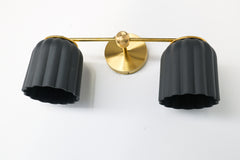 Handmade Double Marcella Sconce in unglazed matte black ceramic — two fluted, scalloped shades on a slim brass arm and round backplate. The bare, unglazed clay gives each shade a soft, stony matte surface with subtle handmade variation, while the Deco-inspired ribbing keeps the silhouette feminine and architectural at once. Black grounds the brass beautifully, so this small-batch ceramic sconce works as a graphic statement or a quiet anchor — flanking a mirror, framing a bed, or lining a hallway. The downwa