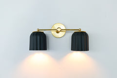 Handmade Double Marcella Sconce in unglazed matte black ceramic — two fluted, scalloped shades on a slim brass arm and round backplate. The bare, unglazed clay gives each shade a soft, stony matte surface with subtle handmade variation, while the Deco-inspired ribbing keeps the silhouette feminine and architectural at once. Black grounds the brass beautifully, so this small-batch ceramic sconce works as a graphic statement or a quiet anchor — flanking a mirror, framing a bed, or lining a hallway. The downwa