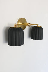 Handmade Double Marcella Sconce in unglazed matte black ceramic — two fluted, scalloped shades on a slim brass arm and round backplate. The bare, unglazed clay gives each shade a soft, stony matte surface with subtle handmade variation, while the Deco-inspired ribbing keeps the silhouette feminine and architectural at once. Black grounds the brass beautifully, so this small-batch ceramic sconce works as a graphic statement or a quiet anchor — flanking a mirror, framing a bed, or lining a hallway. The downwa