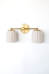 Handmade Double Marcella Sconce in unglazed cream ceramic, seen at a three-quarter angle that shows off the fluted, scalloped silhouette. The bare, unglazed clay gives each shade a soft matte surface with subtle handmade variation, while the Deco-inspired ribbing keeps the shape feminine and architectural. Warm brass ball finials and a round backplate ground the pair. A small-batch ceramic sconce that flatters a mirror, bed, or hallway. Shop handmade ceramic sconces, unglazed cream wall lighting, and fluted
