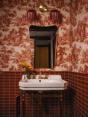 This is the powder room that makes people stop scrolling: a tiny space gone all-in on rusty red, with pastoral toile wallpaper meeting glossy square tile in the same warm terracotta tone. Brass does the heavy lifting throughout, from the console sink legs to the shaped Italian-style mirror to the faucet, and a little yellow and orange bouquet keeps it from feeling too serious. Anchoring the vanity is the Double Marcella sconce, its two ribbed cranberry-red glass shades glowing against the wallpaper and pick