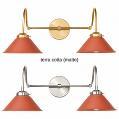 A terracotta two-light gooseneck wall sconce with matte cone shades, shown in Terra Cotta with brass or nickel hardware. It has two curved gooseneck arms joined by a straight crossbar at a round backplate, so it sits wide like a vanity light. Hang it over a bathroom mirror, above a kitchen sink, or over open shelving. Matte clay red sits well with cream plaster, zellige tile, and warm wood, so it suits Mediterranean, Spanish revival, and earthy boho rooms.