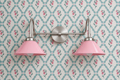 Two-light vanity light with pink gloss cone shades on curved nickel gooseneck arms and a round nickel backplate. Use it over a bathroom mirror or as a double wall sconce above a bed or vanity table. Here it sits on cream wallpaper with pink strawberries inside a teal trellis, so the shades pull the pink from the print. Pink, teal and nickel fit grandmillennial and cottage rooms, a pastel girls bathroom or nursery, and kids bathroom lighting with a sweet vintage feel.