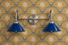 Two-light vanity light with cobalt blue gloss cone shades on curved nickel gooseneck arms and a round nickel backplate. Hang it over a bathroom mirror or use it as a double wall sconce in a kitchen or entry. Here it sits on mustard wallpaper with blue scallops, dots and cream flowers, a block print look. Blue against mustard is a strong contrast that suits eclectic and maximalist rooms, blue bathroom lighting, and a patterned powder room with a global or folk feel.