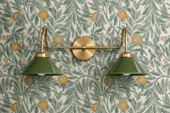 Two-light vanity light with olive green gloss cone shades hanging from curved brass gooseneck arms on a round brass backplate. The shades point down, so it works over a bathroom mirror or as a double wall sconce above a dresser or console. Here it sits on a botanical wallpaper of sage leaves, oranges and pink blossoms on cream. Green and brass suit English cottage, grandmillennial and traditional rooms, and look right in a powder room with patterned walls.