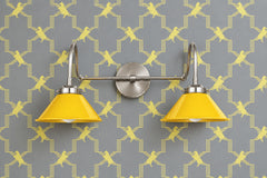 Two-light vanity light with bright yellow gloss cone shades on curved nickel gooseneck arms and a round nickel backplate. Mount it over a bathroom mirror as a vanity light or use it as a double wall sconce in a hallway or kitchen. Here it sits on gray wallpaper with a yellow stepped lattice and birds. The yellow and gray palette with nickel fits art deco bathroom lighting, Hollywood Regency rooms and a bold powder room where the lights match the pattern.