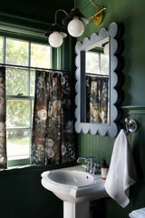 Two-light brass wall sconce with curved arms, black shades, and white glass globes, mounted above a scalloped mirror. The bathroom is painted deep green top to bottom, with beadboard wainscoting and trim in the same color. A powder blue scalloped mirror, dark floral cafe curtains, a white pedestal sink, and chrome faucet mix brass and chrome without fuss. A moody green bathroom idea for an old house or cottage, with vintage-style bathroom vanity lighting.