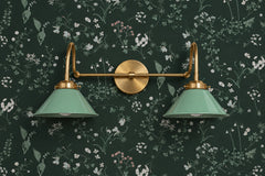 Two-light vanity light with sage green gloss cone shades on curved brass gooseneck arms and a round brass backplate. Hang it over a bathroom mirror or use it as a double wall sconce above a bed or sideboard. Here it sits on dark green wallpaper with small white and pink wildflowers, so the pale green shades read soft against the deep wall. Green on green with brass fits moody bathroom lighting, cottagecore and English country rooms, and a dark powder room.