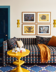 Bright yellow cone-shaped Dryades Sconces add a cheerful pop of color to a maximalist living room with retro influences. The space features a patterned geometric sofa in rust, teal, and navy; a sculptural round yellow side table; and a gallery wall of mystical, folk-art-inspired illustrations. A deep teal painted door and royal blue rug with diamond patterning create bold contrast. This playful and eclectic setup is perfect for customers looking to add modern lighting to a colorful, midcentury-meets-bohemia