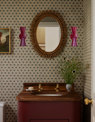 Two glossy black cherry Dryades Sconces flank a vintage rattan mirror in a traditional-meets-whimsical bathroom. The space features neutral peacock-patterned wallpaper, a rich maroon vanity with curved wood top, brass fixtures, and floral decor. The overall look blends traditional European elegance with bold color and playful pattern. A perfect inspiration for shoppers seeking statement sconces to complement jewel-toned accents, warm brass details, and wallpapered bathrooms with a vintage twist.
