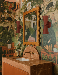 Terracotta-colored Dryades Sconces flank a carved wood scalloped mirror in a dramatic, European-inspired bathroom featuring peach-toned marble, mural-style botanical wallpaper in green, and antique brass fixtures. The pink-brown stone vanity with an arched backsplash ties in with earthy tones throughout the space, while the whimsical, folkloric wallpaper evokes the charm of a storybook. This bathroom is ideal for customers seeking unique wall lighting to pair with warm stone finishes, painterly wallpaper, a