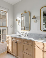 Cream and brass wall sconces in a traditional style bathroom with light wood cabinetry and marble countertops. Mixed with polished nickel sink hardware and brass cabinet knobs and mirrors.  Neutral bathroom has a traditional vibe with modern touches.