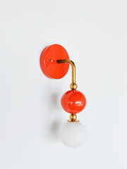 Warm, colorful vanity lighting that gives a bathroom or powder room real personality. Seen in profile, the Ida sconce shows off its curved brass arm reaching from a glossy flame-orange backplate down to a stacked orange sphere and frosted glass globe. The side view highlights the brass ball detailing and how far the fixture projects off the wall. A spirited, saturated choice for eclectic, maximalist, and organic modern interiors.