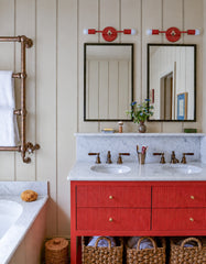 Flame and chrome Thalia Sconces shown as matching horizontal vanity lights above twin mirrors in a bright double sink bathroom with beige paneled walls, a red-orange fluted vanity, white marble counter, bronze plumbing fixtures, copper towel warmer, and woven storage baskets. The slim two-light sconces add a clean modern pop of color while still working with the traditional millwork and marble details. Great for searches like colorful bathroom sconces, red vanity bathroom, double vanity lighting, powder roo