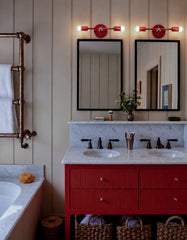 Lit Flame and chrome Thalia Sconces installed horizontally above two black-framed mirrors in a warm, traditional double vanity bathroom. The room features taupe vertical wall paneling, a bold red-orange fluted vanity, white marble countertop and backsplash, copper towel warmer, antique-style plumbing, wicker baskets, and moody old-house charm. A strong example of colorful bathroom lighting for double vanities, red bathroom vanities, European-inspired bathrooms, traditional interiors, and playful modern scon