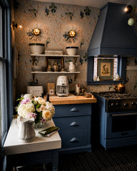 Moody blue kitchen with a statement range hood, open shelves styled with ceramics, and delicate floral wallpaper for an English cottage, vintage-inspired feel. Warm brass lighting adds contrast and polish against the soft blue palette. Great for blue kitchen ideas, wallpapered kitchen, cottage kitchen decor, brass lighting, and statement ceiling fixtures.