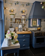 Moody blue kitchen with a statement range hood, open shelves styled with ceramics, and delicate floral wallpaper for an English cottage, vintage-inspired feel. Warm brass lighting adds contrast and polish against the soft blue palette. Great for blue kitchen ideas, wallpapered kitchen, cottage kitchen decor, brass lighting, and statement ceiling fixtures.