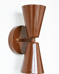 Chocolate brown is having a moment in moody, sophisticated interiors, and this double-cone Dryades sconce leans right into it. The bowtie silhouette is pure mid-century modern, finished in a glossy Fudge Brown powder coat with warm brass hardware at the center collar and mounting screws. The two opposing cones throw light up and down, making it a smart pick for flanking a bathroom mirror, a bedside, or a hallway that needs a little richness. A confident color choice for anyone building a collected, anti-bei