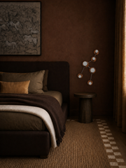 A moody, masculine bedroom with a rich monochromatic brown palette, featuring a dark upholstered bed, layered neutral and chocolate bedding, a hand-hewn or plaster-look wall finish, dramatic drapery, and a sculptural side table. Mounted beside the bed is the Taurus sconce by Sazerac Stitches, a constellation-inspired wall light with circular shades and a celestial branching form. The sconce is shown in a warm earthy colorway that blends into the room’s color-drenched design, making it ideal inspiration for 