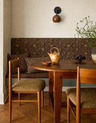 Warm, cozy breakfast nook featuring a built-in banquette with patterned upholstery, layered neutral pillows, and a round wood pedestal table with vintage-style dining chairs on herringbone hardwood floors. The Loa wall sconce in glossy fudge brown with matte black hardware adds sculptural, modern mid-century style and targeted downlight, perfect for small dining spaces, banquette seating, café corners, and breakfast nook lighting ideas. Great search terms: modern wall sconce, mid century modern lighting, br