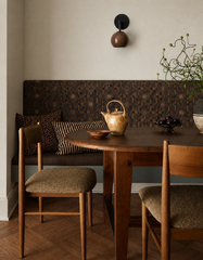 Cozy breakfast nook and dining banquette styled in warm neutrals, featuring a built-in upholstered bench with vintage-inspired geometric fabric, a round wood pedestal table, and wood dining chairs on herringbone flooring. A sculptural Loa wall sconce with a glossy fudge-brown dome shade and matte black backplate adds soft ambient light and a modern mid century accent. Great inspiration for banquette lighting, breakfast nook wall lighting, organic modern decor, and small dining space design.