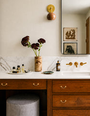 Warm neutral bathroom vanity styled with a glossy fudge-brown Loa wall sconce and aged brass backplate, mounted above a white marble countertop with brass faucet and walnut wood drawers. A minimalist black-framed mirror, perfume bottles, and a carved vase with dark blooms create a modern traditional, organic modern look. Ideal inspiration for bathroom vanity lighting, powder room sconces, mid century modern brass sconce lighting, spa bathroom decor, and warm minimalist interiors.