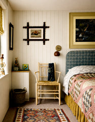 Fudge brown dome Loa wall sconce with polished chrome backplate and arm, mounted on a white beadboard wall in a cozy eclectic vintage bedroom. Styled as a bedside reading light beside a blue plaid upholstered headboard and layered patchwork quilt, with a woven wood chair, framed folk art, and a colorful patterned rug. Warm, collected cottage style with mid century modern accents, perfect for bedroom wall lighting, bedside sconces, and small-space ambient lighting.