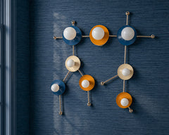 Gemini One wall sconce in a denim blue, mustard yellow, and cream color palette with polished metal hardware, mounted on rich dark blue grasscloth wallpaper. Inspired by the Gemini star constellation, this sculptural wall light combines playful color with a clean, graphic silhouette that feels both artistic and tailored. The textured wallpaper adds depth and a refined designer look, making this a strong choice for eclectic interiors, colorful powder rooms, kids spaces, playrooms, reading nooks, or any room 