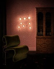 Dramatic evening view of a peach Gemini One sconce glowing against pink moire wallpaper in a dark, vintage-inspired lounge. The constellation wall light adds warm sculptural lighting above an olive green velvet chair, small cocktail table, patterned rug, and burl wood cabinet. This image blends moody maximalist decor, zodiac lighting, celestial wall art, colorful sconces, retro glam style, and cozy cocktail room inspiration.
