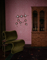 Moody maximalist lounge with pink moire wallpaper, an olive green velvet sculptural chair, dark floral rug, and a burl wood bar cabinet styled with wine bottles and glassware. The peach Gemini One sconce sits on the wall like celestial art, with round star discs, white globe bulbs, and a constellation-inspired shape based on the Gemini zodiac sign. A strong fit for eclectic interiors, astrology decor, colorful statement lighting, vintage glam rooms, and artful wall sconces.