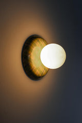 Lit close angled view of a glossy lime green and pale blue plaid ceramic wall sconce with a matte white globe shade, showing warm ambient light and a soft halo on the wall, a colorful statement globe sconce for maximalist interiors, dopamine decor, playful modern lighting, and artful accent lighting in a hallway, bedroom, or powder room.