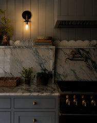 Moody grey kitchen with the black and silver Adele sconce turned on, casting a warm glow over vertical wall paneling, stacked books, glassware, and a green-veined marble counter. The lit wall sconce brings soft ambient light to a richly layered kitchen with grey cabinetry, marble backsplash, scalloped stone detail, brass pot filler, and professional range. A strong image for modern traditional kitchen lighting, black wall sconces, silver accent lighting, marble kitchen ideas, and cozy evening kitchen stylin