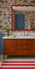 Playful wallpapered bathroom with blue wainscoting, blue door trim, a red wavy mirror, marble counter, wood vanity, brass faucet, and a red-and-cream striped rug. The Hydrangea Blue Medium Thalia Sconce is mounted vertically beside the mirror, with a blue round backplate, slim bar, brass accents, and white globe bulbs. The space mixes cottagecore, vintage modern, and colorful maximalist bathroom style.
