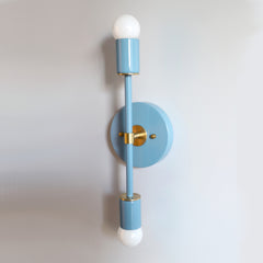 Pastel blue and brass Medium Thalia Sconce with double globe bulbs, soft color-drenched mid century modern wall sconce with a sleek vertical silhouette and round backplate, modern coastal and Scandinavian-inspired lighting for bathroom vanity, bedside walls, nursery decor, or hallway accents by Sazerac Stitches