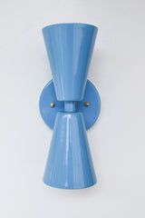 Close-up image of the Dryades Sconce in a glossy hydrangea blue powder-coated finish, featuring a mid-century modern double-cone silhouette with a matching blue backplate and subtle brass hardware accents. This vibrant wall light fixture is handcrafted with clean geometric lines and a bold color that adds a playful, sculptural element to contemporary interiors. Ideal for use in powder rooms, bedrooms, hallways, or as statement lighting in eclectic and maximalist homes. Shown against a neutral white backgrou