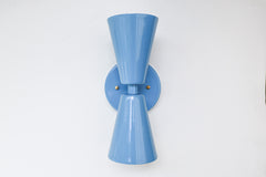 Close-up image of the Dryades Sconce in a glossy hydrangea blue powder-coated finish, featuring a mid-century modern double-cone silhouette with a matching blue backplate and subtle brass hardware accents. This vibrant wall light fixture is handcrafted with clean geometric lines and a bold color that adds a playful, sculptural element to contemporary interiors. Ideal for use in powder rooms, bedrooms, hallways, or as statement lighting in eclectic and maximalist homes. Shown against a neutral white backgrou