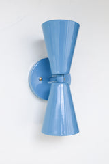 Close-up image of the Dryades Sconce in a glossy hydrangea blue powder-coated finish, featuring a mid-century modern double-cone silhouette with a matching blue backplate and subtle brass hardware accents. This vibrant wall light fixture is handcrafted with clean geometric lines and a bold color that adds a playful, sculptural element to contemporary interiors. Ideal for use in powder rooms, bedrooms, hallways, or as statement lighting in eclectic and maximalist homes. Shown against a neutral white backgrou