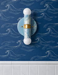 Hydrangea, Cream, and Gold hand-marbled Small Thalia Sconce installed vertically on dark blue wave wallpaper above glossy white bathroom tile. The soft blue and cream marbling pairs with the nautical wallpaper for a coastal bathroom, beach house powder room, kids bath, or playful blue and white guest bathroom. The compact two-light design, brass hardware, and globe bulbs make it a fun decorative wall sconce for small bathrooms, patterned walls, and colorful interiors.