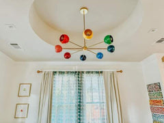 Arcenciel chandelier in bright rainbow colorway with glossy multicolor globe shades on a warm brass sputnik frame, hanging from a long downrod inside a recessed tray ceiling above sheer curtains and a large window, a bold mid century modern statement chandelier for maximalist and eclectic interiors, perfect as modern dining room lighting, living room ceiling light, or playful family room centerpiece.