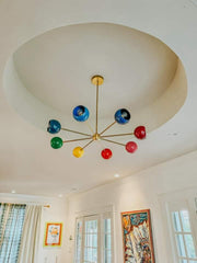 Close view of the Arcenciel rainbow sputnik chandelier featuring bright red, blue, green, yellow, and coral globe shades on branching brass arms, installed in a white room with a dramatic tray ceiling and colorful art, a modern mid century inspired statement ceiling light for design-forward homes, ideal for dining rooms, living rooms, entryways, and contemporary eclectic spaces that love bold color.