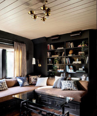 Cozy living room with a warm, inviting atmosphere featuring a built-in corner sofa with brown cushions and patterned pillows. The room has dark walls and a wooden ceiling with a brass chandelier. A large bookshelf filled with books and decor items lines the back wall, adding a touch of sophistication. Natural light streams in through a nearby window, illuminating the space and highlighting the glass coffee table in the center.  Libra constellation shaped ceiling light by Sazerac Stitches.