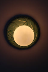 Jade green ceramic wall sconce lit, warm glow through frosted globe, glossy leaf relief backplate, chinoiserie botanical accent light with soft halo